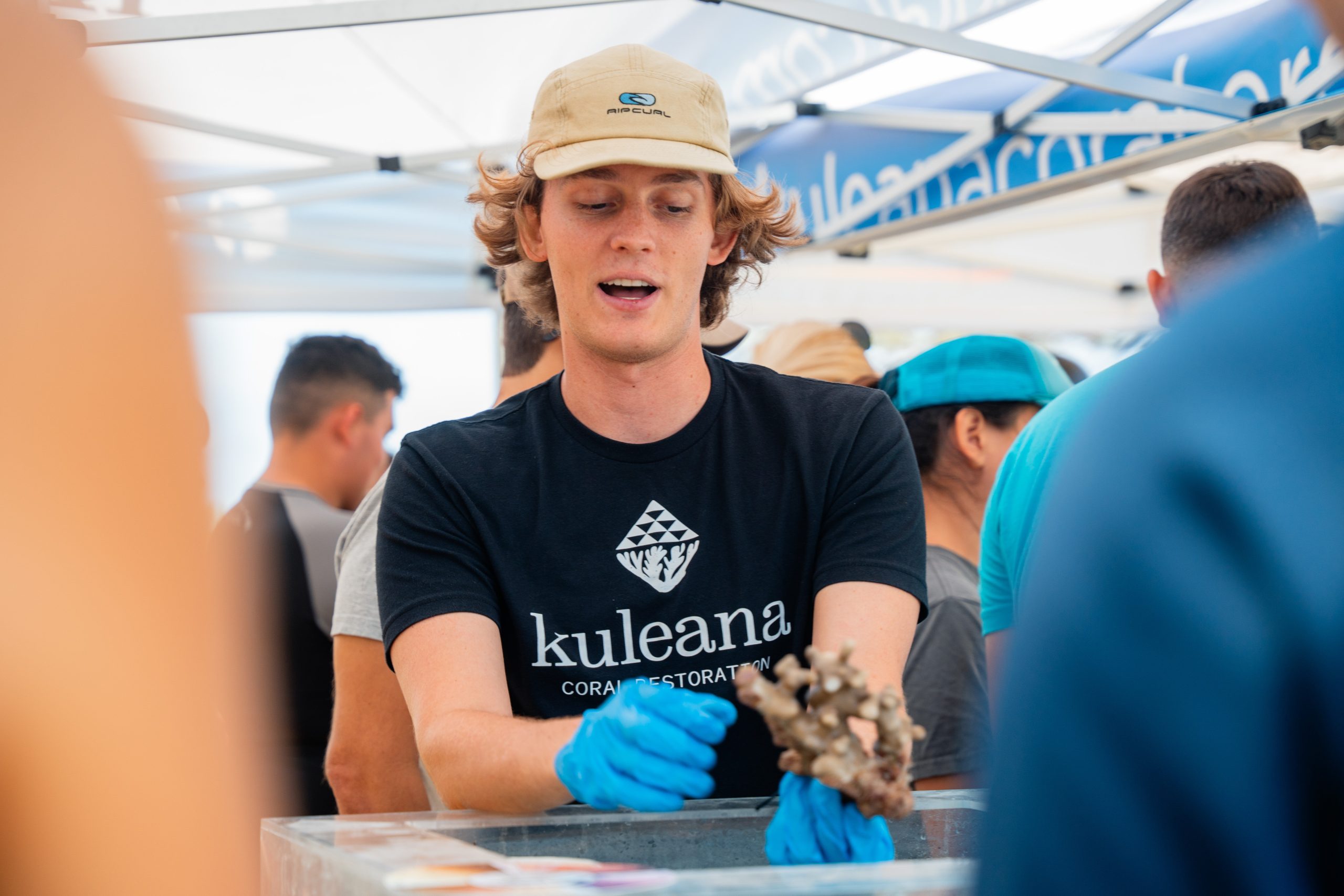 A man wearing a hat and blue gloves holds a piece of fish, showcasing his catch with a focused expression.